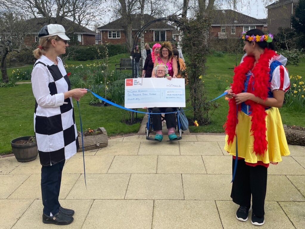 To members of staff in fancy dress hold a blue ribbon between them. About to pass through the ribbon is a member of staff, pushing a resident in  a wheelchair. The resident is holding a large cheque.