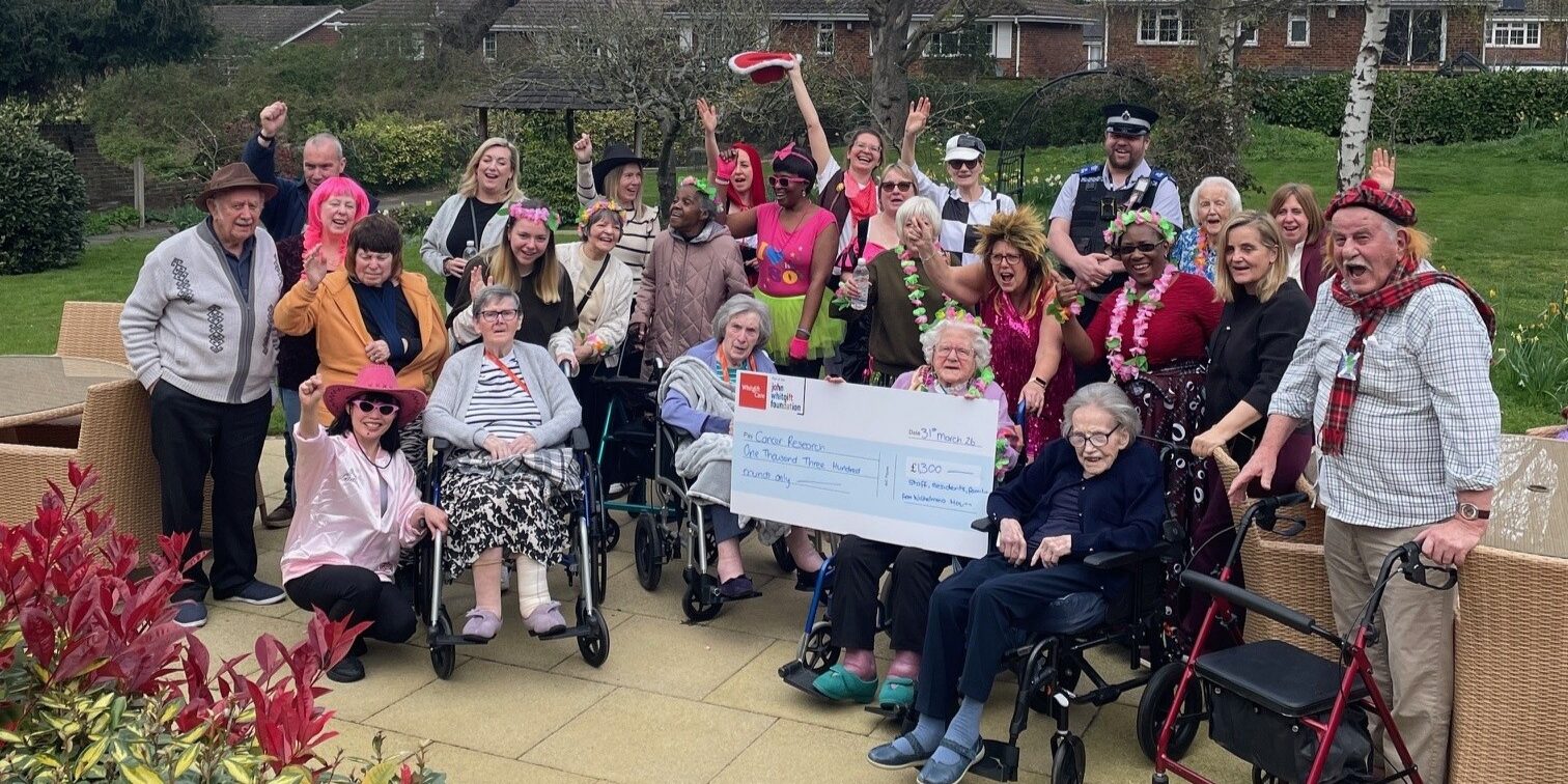 A group of men and women, some in brightly coloured fancy dress, pose for a group photo in the garden at Wilhelmina House