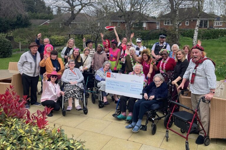 A group of men and women, some in brightly coloured fancy dress, pose for a group photo in the garden at Wilhelmina House