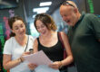 A man and woman look over the shoulder of a female student as she opens her A-Level results. They all look delighted.