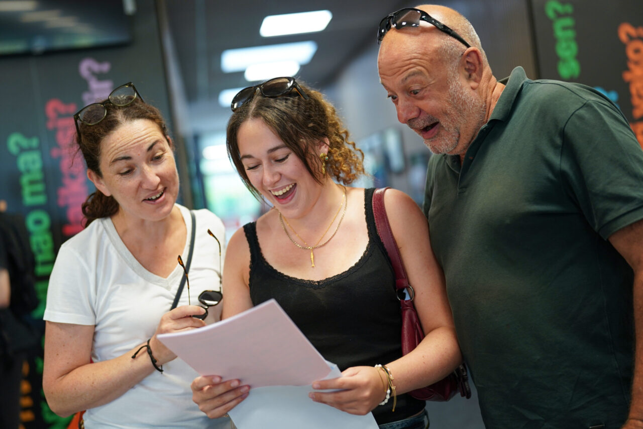A man and woman look over the shoulder of a female student as she opens her A-Level results. They all look delighted.