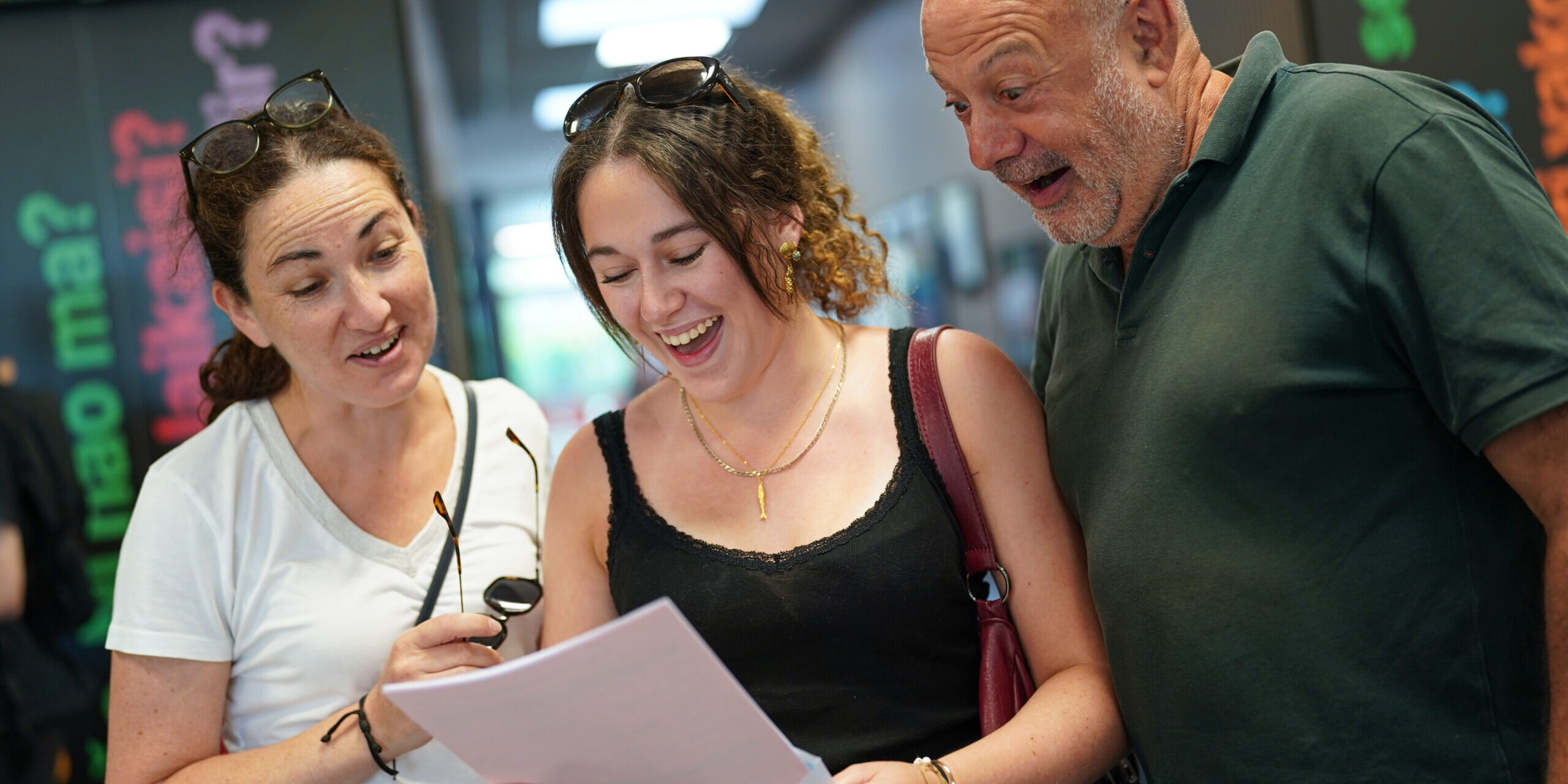 A man and woman look over the shoulder of a female student as she opens her A-Level results. They all look delighted.