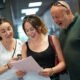 A man and woman look over the shoulder of a female student as she opens her A-Level results. They all look delighted.