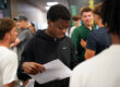 A student wearing a dark sweatshirt looks at his GSCE results while other students are visible in the background.