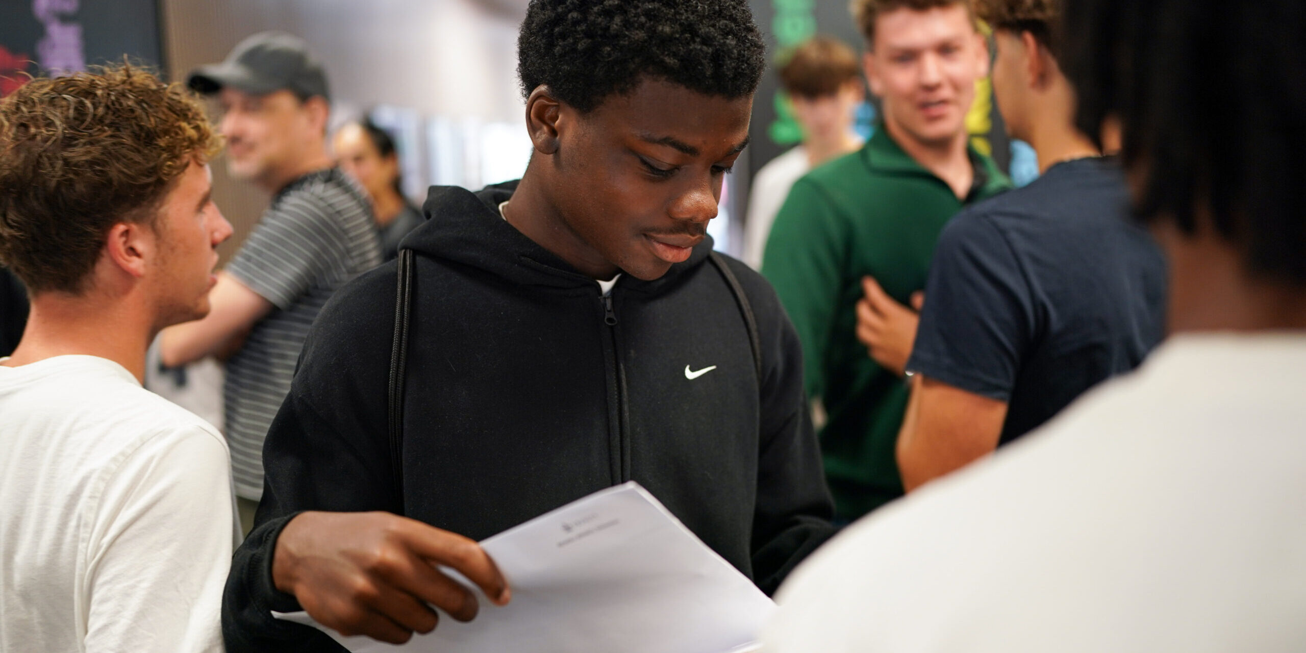 A student wearing a dark sweatshirt looks at his GSCE results while other students are visible in the background.