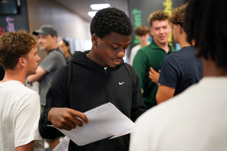 DF201349 A student wearing a dark sweatshirt looks at his GSCE results while other students are visible in the background.